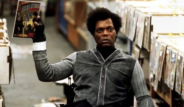 A man (Elijah Price from the movie unbreakable) with short curly hair and a serious expression sits in a wheelchair inside a comic book store. He wears a dark velvet jacket with light gray sleeves and holds up a comic book in one hand while surrounded by shelves filled with colorful comic issues.