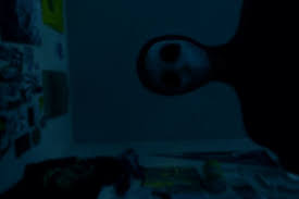 A dimly lit, blue-tinted photograph showing a humanoid figure with a pale, featureless face and dark, hollow eyes leaning over the camera. The figure appears to be wearing a black hood or bodysuit, its head tilted slightly downward as if peering at the viewer. The background shows the corner of a cluttered room with posters and papers on the walls, but the lighting is so dark that details are obscured. The eerie composition and cold blue tones create a sense of dread and intrusion, as though the figure is silently watching or approaching.