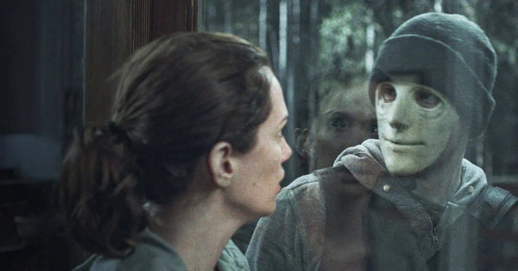 A tense scene from the film “Hush.” A woman with her hair tied back stands inside a house, staring through a glass door at a masked man outside. His pale mask and dark beanie create an eerie contrast against the forest behind him, while her reflection in the glass heightens the sense of fear and confrontation.