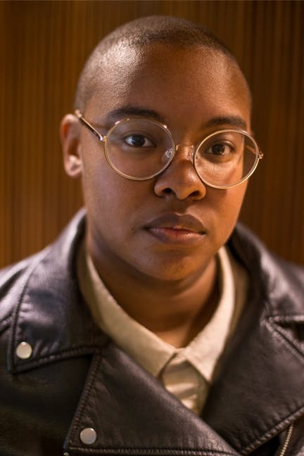 A portrait of Rivers Solomon. They have a shaved head, wear round glasses with thin metal frames, and are dressed in a light collared shirt under a black leather jacket. The background is softly blurred with warm brown tones, drawing focus to their thoughtful gaze and composed presence.
