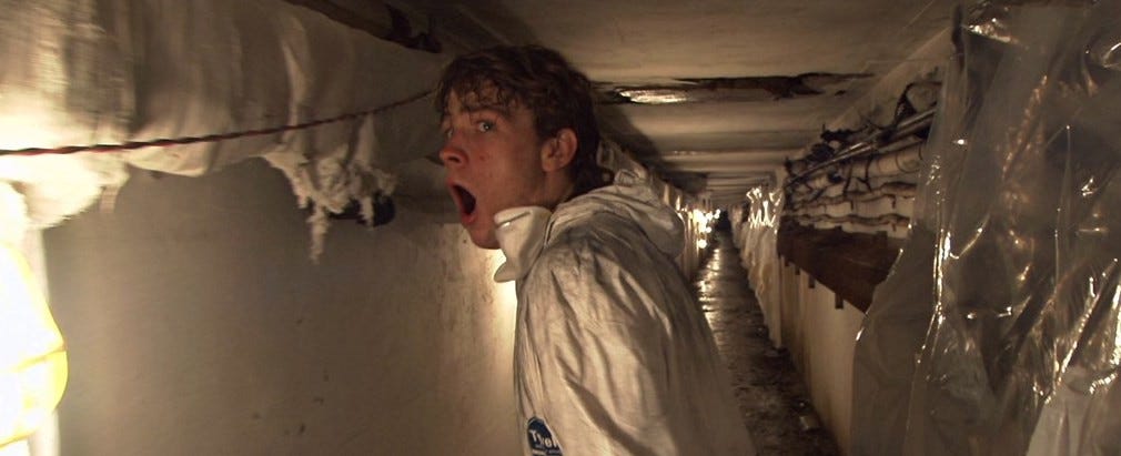 A young man in a white hazmat suit stands in a narrow, dimly lit underground tunnel lined with pipes and plastic sheeting. He turns suddenly, mouth open in shock or fear, as if reacting to something behind him.