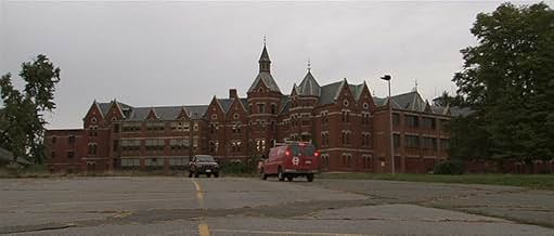 A wide shot of a large, decaying red-brick asylum with Gothic architecture, tall windows, spires, and a central tower, under an overcast sky. Two vehicles are parked in the cracked, empty lot in front of the building.