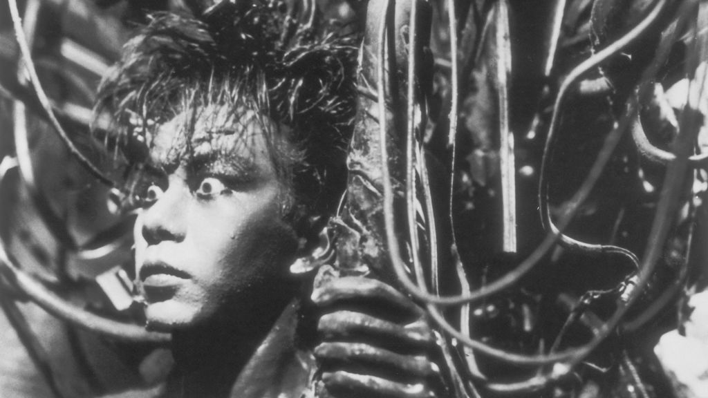 A black-and-white close-up still from the film Tetsuo: The Iron Man. A man’s face, slick with sweat and oil, stares wide-eyed into the distance with an expression of shock or terror. His hair is wild and tangled, and his skin appears fused with metal wires and tubes that coil around his head and shoulders. One hand, darkened and rough as if coated in metal, grips a large cable in the foreground. The scene is dimly lit, with harsh contrasts of light and shadow, creating a claustrophobic and industrial atmosphere.
