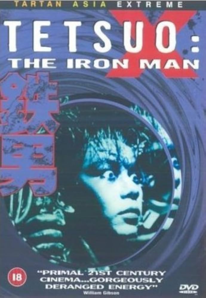 DVD cover for the Japanese cyberpunk horror film Tetsuo: The Iron Man. The background is metallic blue with swirling patterns. At the centre is a man’s face lit in harsh blue light, appearing fused with wires and metal. The title “TETSUO: THE IRON MAN” is printed in bold white and red letters at the top, with large red Japanese characters on the left. A quote at the bottom reads, “Primal 21st century cinema… gorgeously deranged energy” — William Gibson. The cover carries an 18-age rating and a DVD logo.