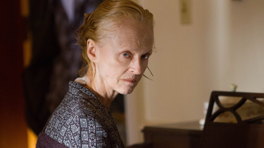 A scene from “The Taking of Deborah Logan.” An older woman with pale skin and thinning hair sits beside a piano, turning to look over her shoulder with a sharp, penetrating gaze. She wears a patterned robe and a light-colored blouse. The lighting is warm but dim, casting soft shadows that emphasize her gaunt features and the tension in her expression. Her posture and expression suggest both suspicion and distress.