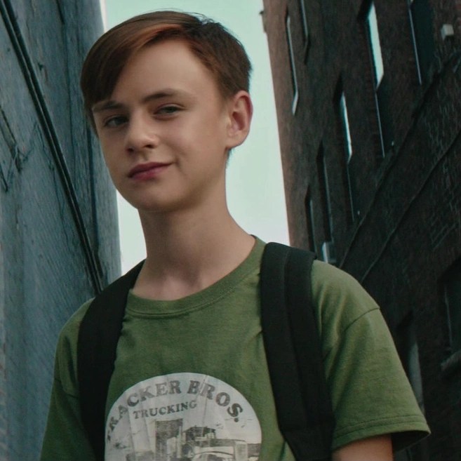 A young white boy (Bill Denbrough) with short brown hair stands in an alleyway between two brick buildings. He wears a green T-shirt and a backpack, looking slightly to the side with a faint, knowing smile. The image is from the film It (2017), depicting the character Bill Denbrough as a child.