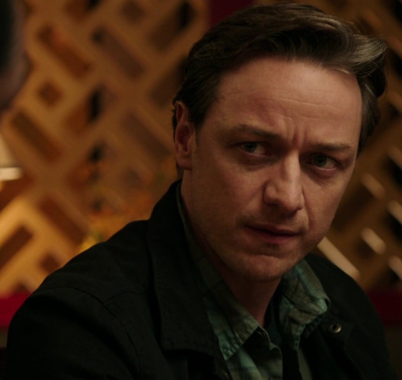 A white man (Bill Denbrough) with short brown hair and a serious expression sits indoors, lit warmly against a wooden lattice background. He wears a green checked shirt and dark jacket. The image is from It Chapter Two (2019), showing the adult version of Bill Denbrough.