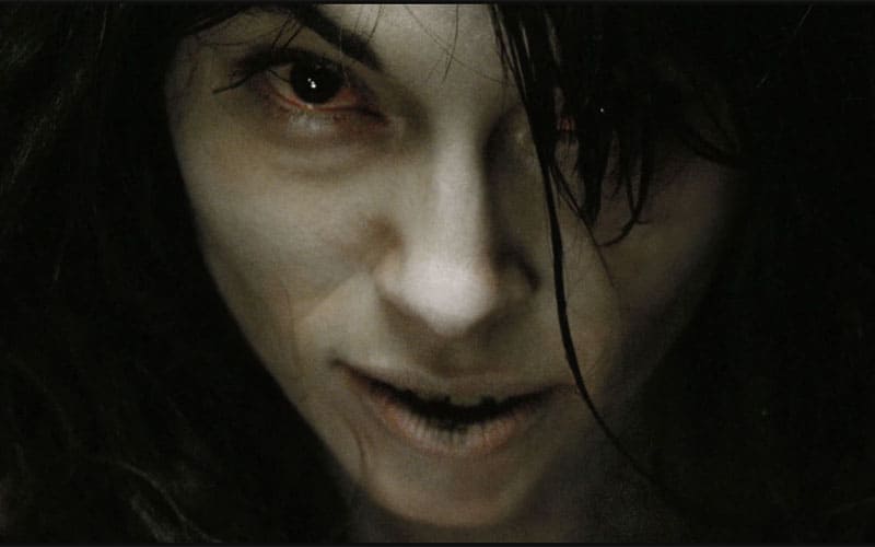 A tight close-up of the same pale young woman from the film, staring directly into the camera with a predatory expression. Her eyes appear red and sunken, her lips are slightly parted, and her dark hair hangs messily around her face. The lighting emphasizes her cold, lifeless skin and the unsettling intensity of her gaze.