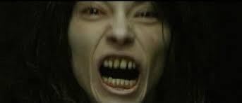 A close-up of a young woman’s face lit dimly, her skin pale and corpse-like. Her mouth is wide open in a scream, revealing darkened, rotten-looking teeth. Her eyes are shadowed, and her long dark hair frames her face, heightening the sense of aggression and horror.