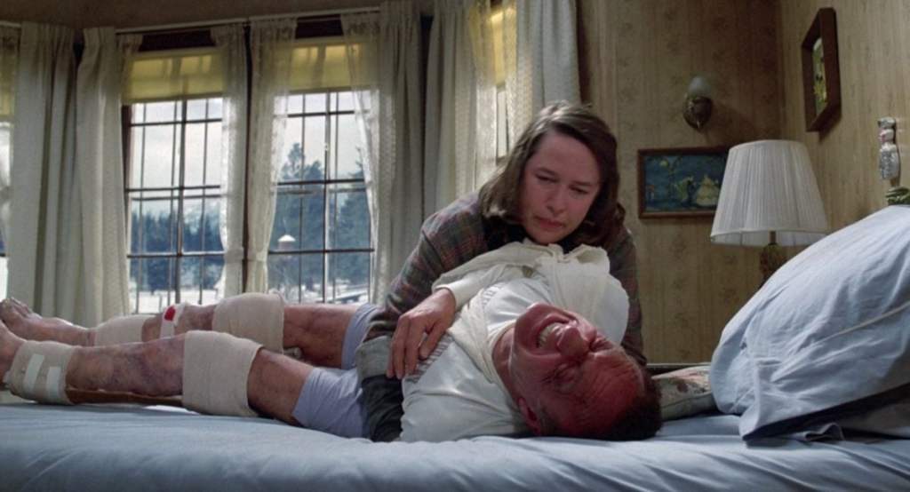 A terrified, injured man lies strapped to a bed in a rustic bedroom with pale floral curtains and large windows showing a snowy landscape outside. His legs are heavily bruised and bandaged, stretched out straight. A woman in a plaid shirt and cardigan leans over him, holding his torso down as he screams in pain. The atmosphere is tense and claustrophobic.