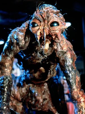 A grotesque humanoid creature with mottled, reddish-brown, decaying skin crouches toward the camera. Its large, glossy black eyes, distorted facial structure, and insect-like features give it a disturbing, partially transformed appearance. The lighting is dim, highlighting wet, organic textures across its body.