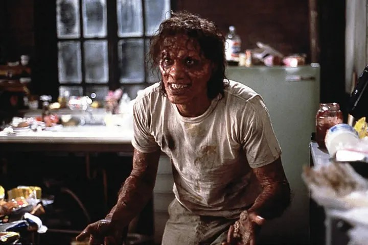 A man stands in a cluttered, dimly lit kitchen, his skin mottled, raw, and partially transformed with insect-like textures. He wears a dirty white T-shirt and beige trousers. His posture is slightly hunched, his hands tense, and his expression strained, conveying pain and fear as the transformation progresses.