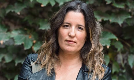 Colour photograph of a woman, Agustina Bazterrica, facing the camera. She has shoulder-length wavy hair with darker roots and lighter ends, and a neutral, calm expression. She is wearing a black leather jacket over a dark top. The background consists of green foliage, softly out of focus.