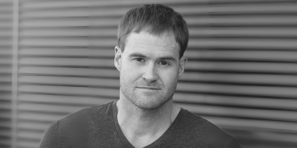 Black-and-white photograph of a man, Craig Davidson, looking directly at the camera. He has short dark hair, light stubble, and a neutral expression. He is wearing a dark V-neck T-shirt. The background consists of horizontal metal or wooden slats, slightly out of focus.