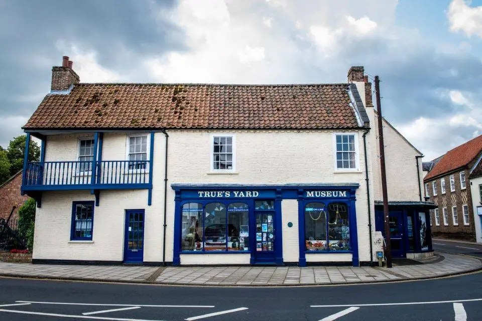 Exterior view of True’s Yard Fisherfolk Museum. A long, low, whitewashed brick building with a red tiled roof stands on a street corner. The ground floor has deep blue painted shopfront windows with the words ‘TRUE’S YARD’ and ‘MUSEUM’ in white lettering. A small wooden balcony painted blue runs along the upper floor on the left side. The sky above is partly cloudy.