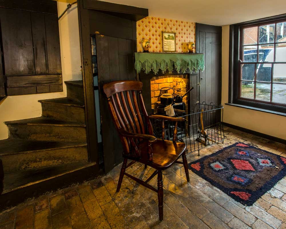 Interior of a historic fisherfolk cottage room. A wooden chair sits beside a lit brick fireplace with a hanging kettle. The floor is made of worn stone tiles, partly covered by a patterned rug. A narrow wooden staircase rises steeply on the left. Dark wooden cupboards line the walls, and a window on the right lets in daylight from a small yard outside.