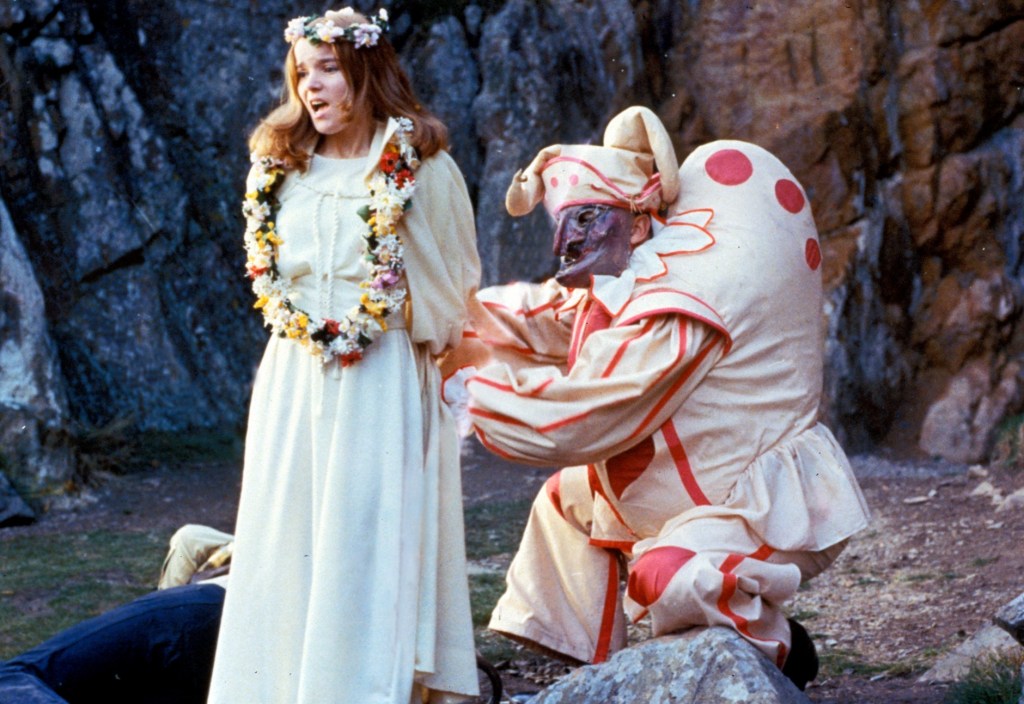 A scene set outdoors among rocky cliffs. A young woman wearing a white dress and a flower crown stands in the foreground, looking distressed. She is restrained by a figure in an elaborate, padded costume resembling a grotesque hunchback clown, decorated with pink dots and a mask over his face. The contrast between the pastoral setting, the woman’s ceremonial clothing, and the unsettling figure creates a sense of ritualistic threat.